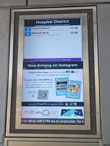 A real-time information screen at the Hospital District bus stop shows upcoming bus arrival times and a RIPTA social media promo.